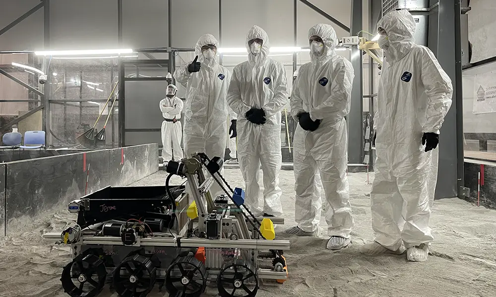 四名穿着实验室清洁服的学生聚集在地面上的一个多轮机器人周围.
